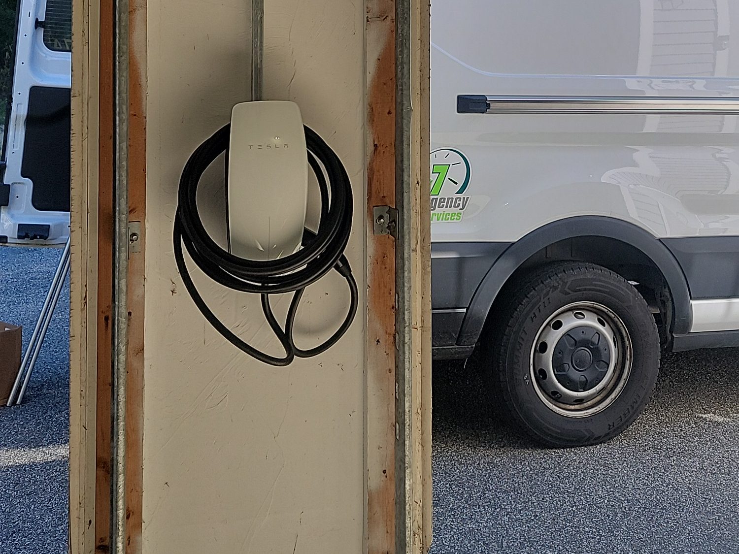 Level 2 wall connector with its cable coiled, mounted on a garage door column and fed by conduit from above, with a work van parked outside