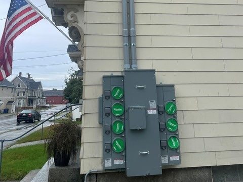 New multi-position meter bank with green meter socket covers and two conduit risers on the side of a clapboard building